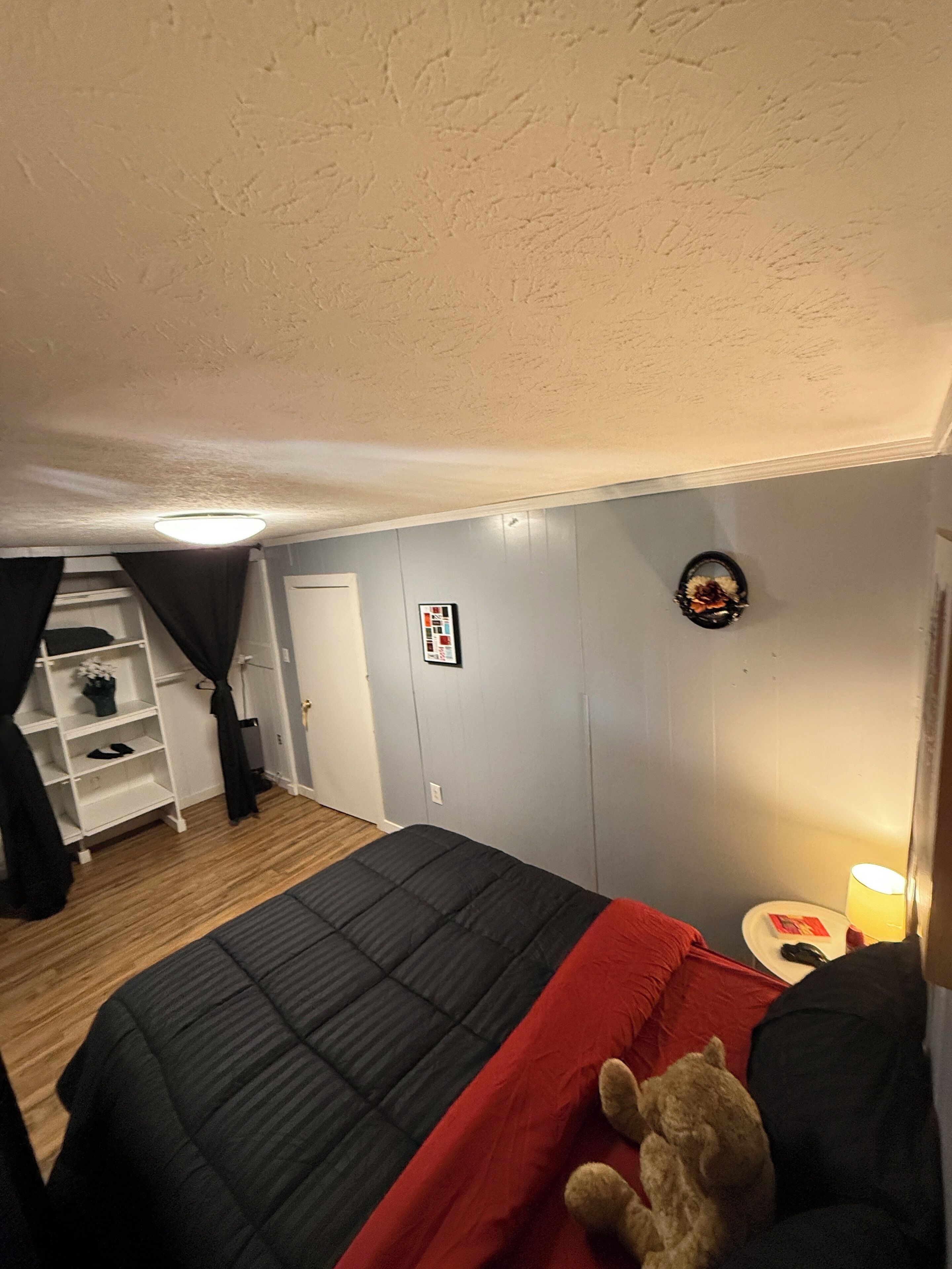 Une chambre à coucher avec un lit, une table de chevet, un ours en peluche, une étagère à chaussures et une porte.