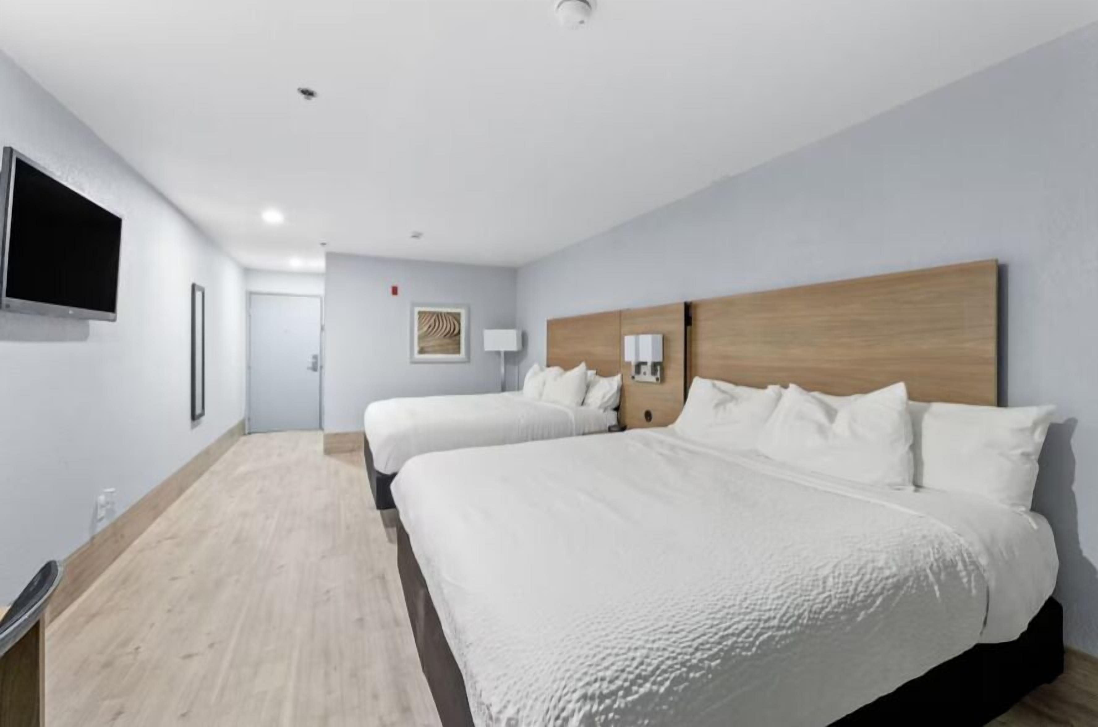 A hotel room with two beds, a flat-screen TV, a wooden headboard, and a small bedside table with a lamp.