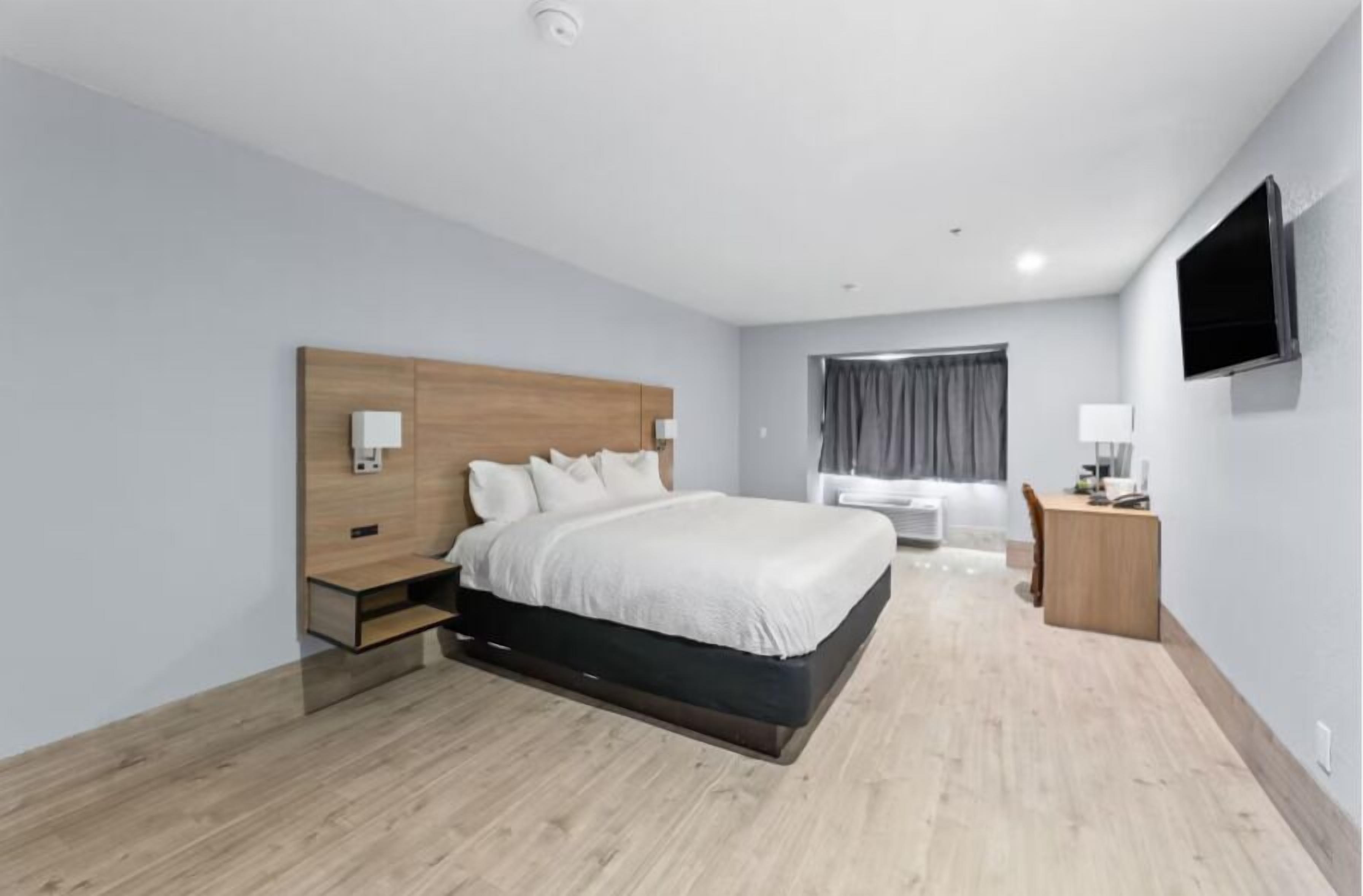A modern hotel room with a wooden headboard, a large bed, a desk, and a flat-screen TV.