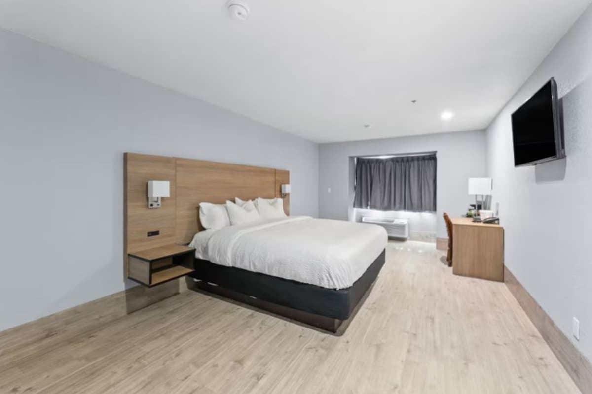 A modern hotel room with a wooden headboard, a large bed, a desk, and a flat-screen TV.