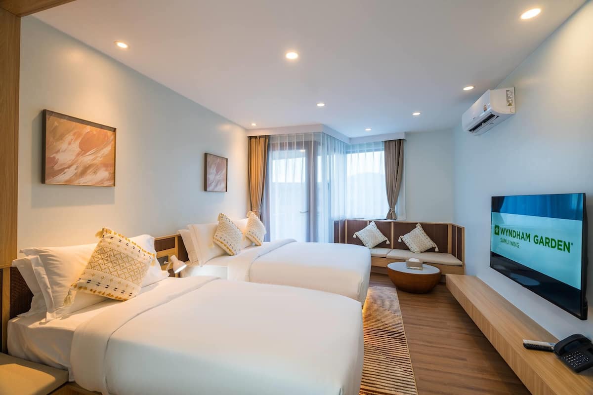 Habitación de hotel con dos camas, un sofá, un televisor y un ventanal grande con cortinas.
