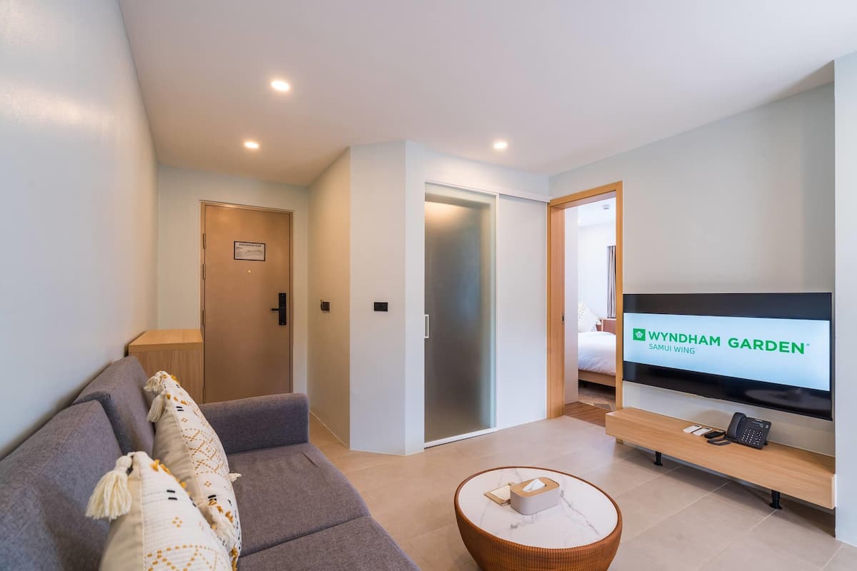 Una sala moderna con un sofá gris, un televisor de pantalla plana, una mesa de centro de madera y un ventanal corredero que da a un dormitorio.