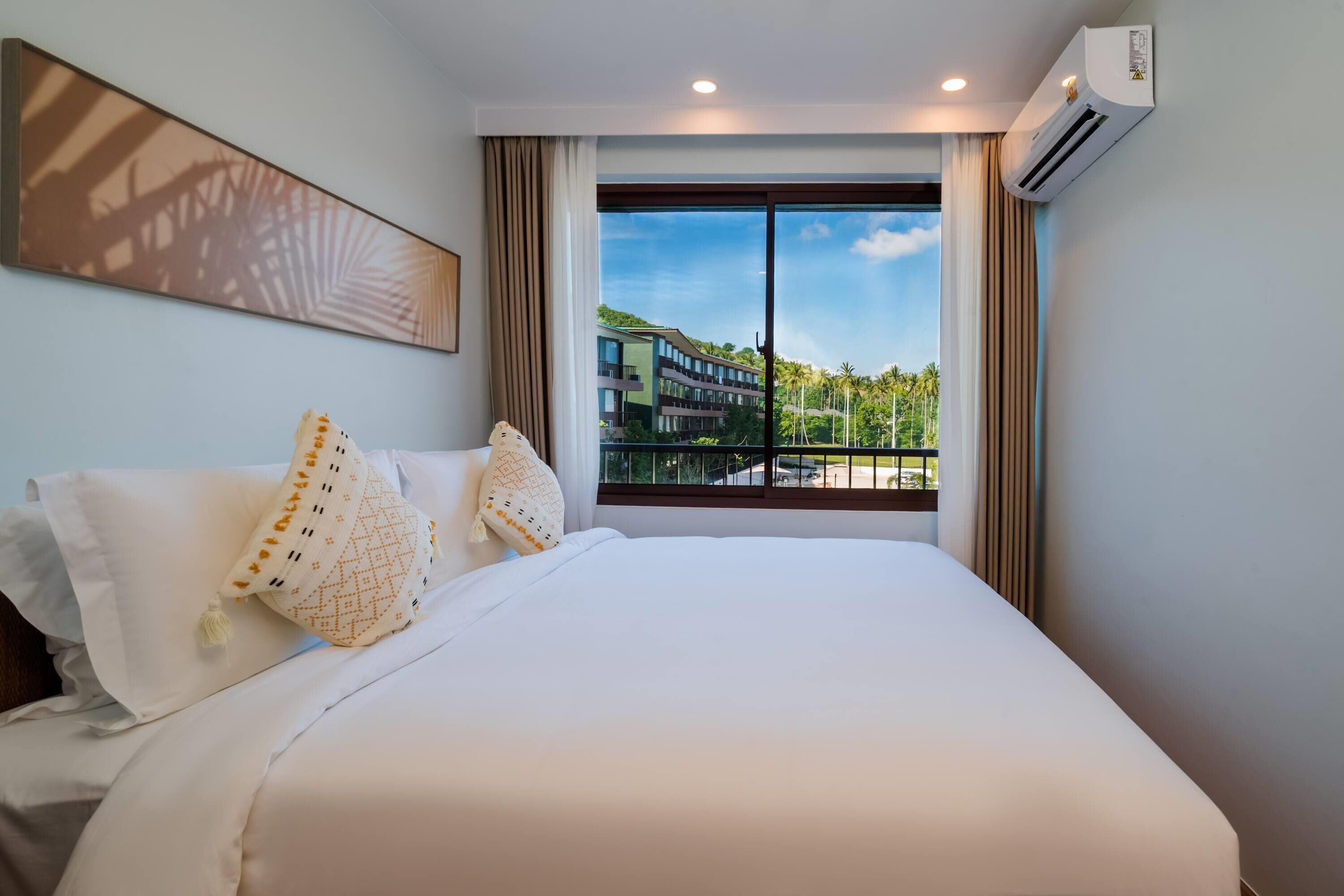 A hotel room with a large bed, two pillows, a wall-mounted air conditioner, and a window with a view of buildings and greenery.