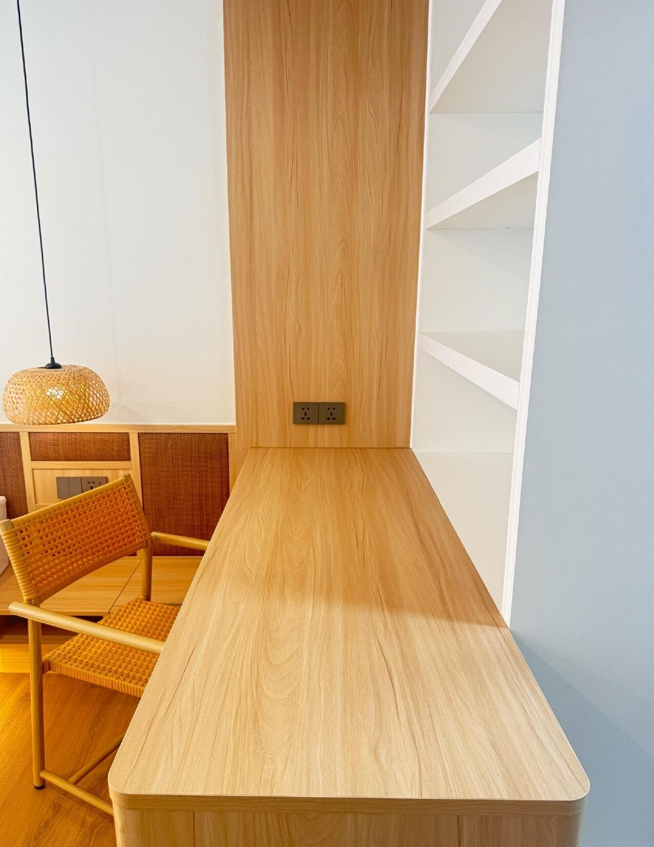 Una mesa de madera sencilla con una silla, una lámpara de fibras tejidas y un enchufe en la pared.