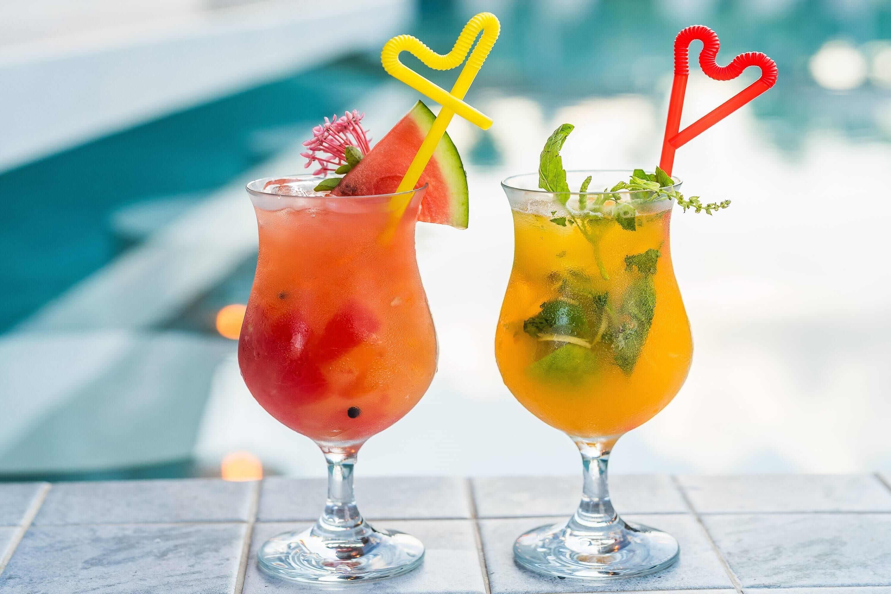 Kaksi sydämenmuotoisilla pilleillä varustettua cocktailia, toisessa vesimeloninviipale ja toisessa mintunlehtiä, laatoitetulla pinnalla uima-altaan läheisyydessä.