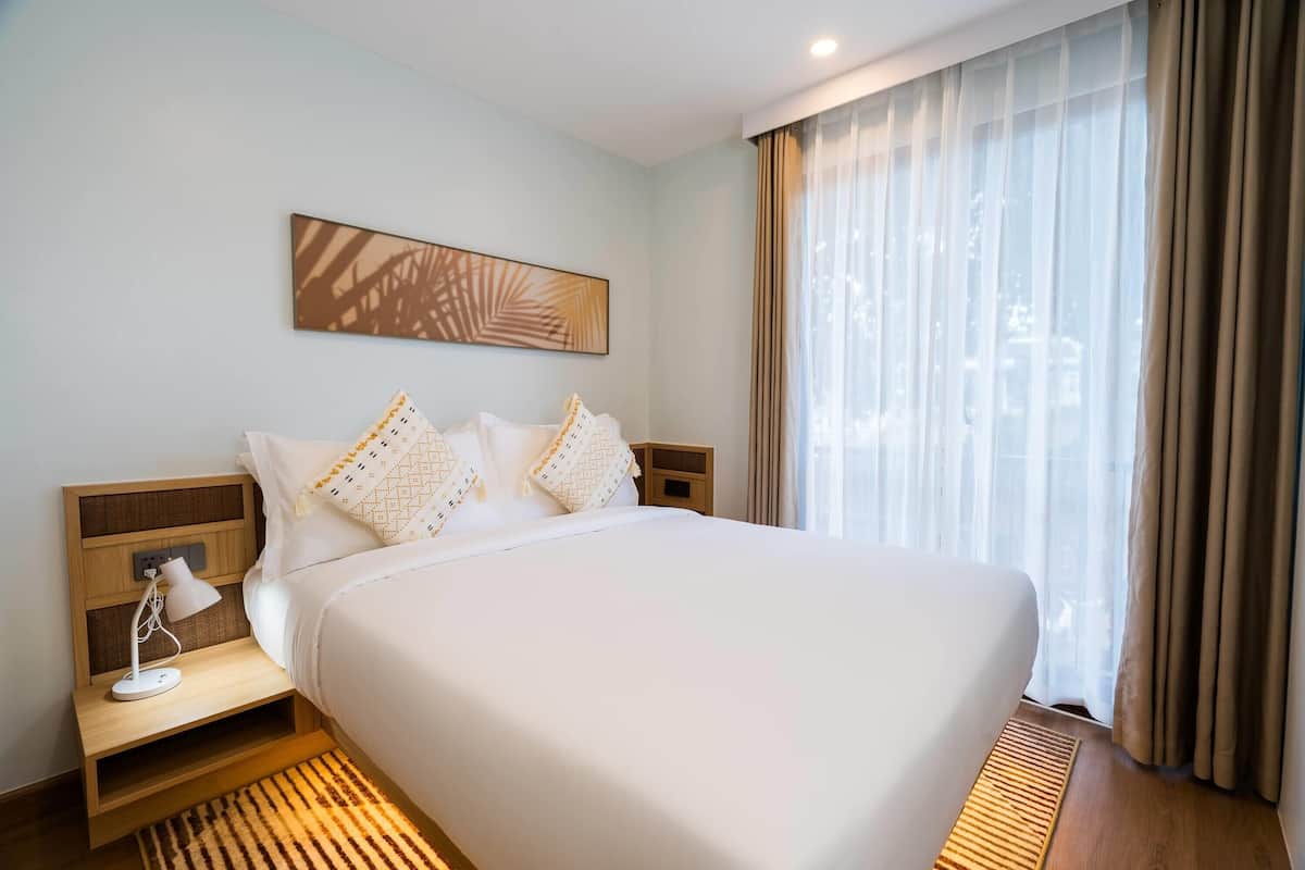 Una habitación de hotel ordenada con una cama grande, una mesita de noche con lámpara, un pequeño tocador y un ventanal con cortinas translúcidas.