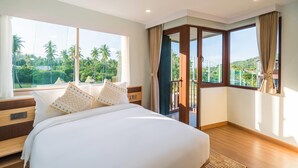 A hotel room with a large bed, a wooden nightstand, a window with a view of palm trees, and a sliding glass door to a balcony. - Wyndham Garden Samui Wing (Koh Samui)