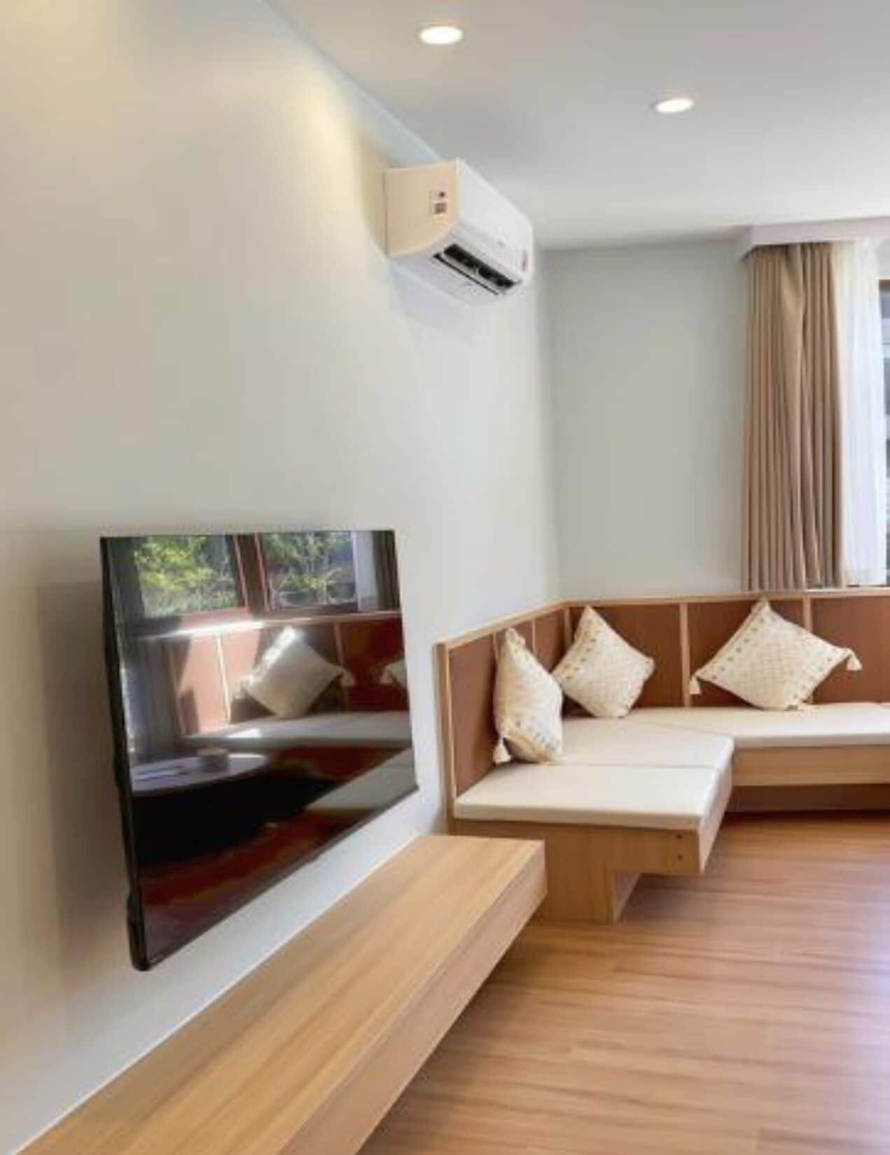 A modern living room with a flat-screen TV mounted on the wall, a wooden bench with cushions, and an air conditioning unit on the ceiling.