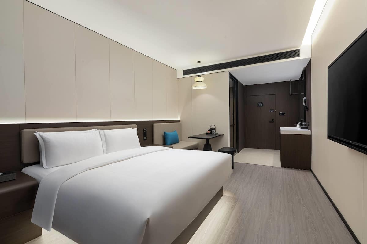 Een moderne hotelkamer met een groot bed, een klein tafeltje met een lamp, een televisie en een badkamer met een wastafel en spiegel.