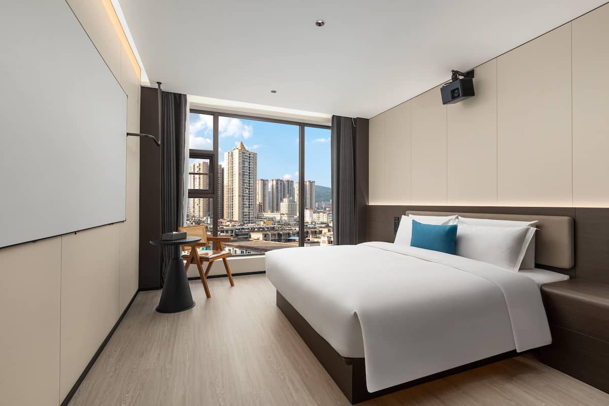 Een moderne hotelkamer met een groot bed, een kleine tafel en een stoel. Grote ramen bieden uitzicht over de stad.