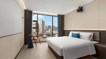 Una habitación de hotel moderna con una cama grande, una mesita y una silla. Se aprecia vista panorámica de la ciudad a través de amplios ventanales.
