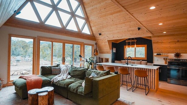 A cozy cabin interior with a large wooden ceiling, a dining area with bar stools, a kitchen with a stove, and a view through large windows. - Hello Dreamer A-Frame (Maysville)