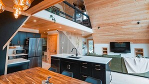 A modern kitchen with a central island, a dining area with a wooden table, and a living area with a sofa and TV. - Hello Dreamer A-Frame (Maysville)