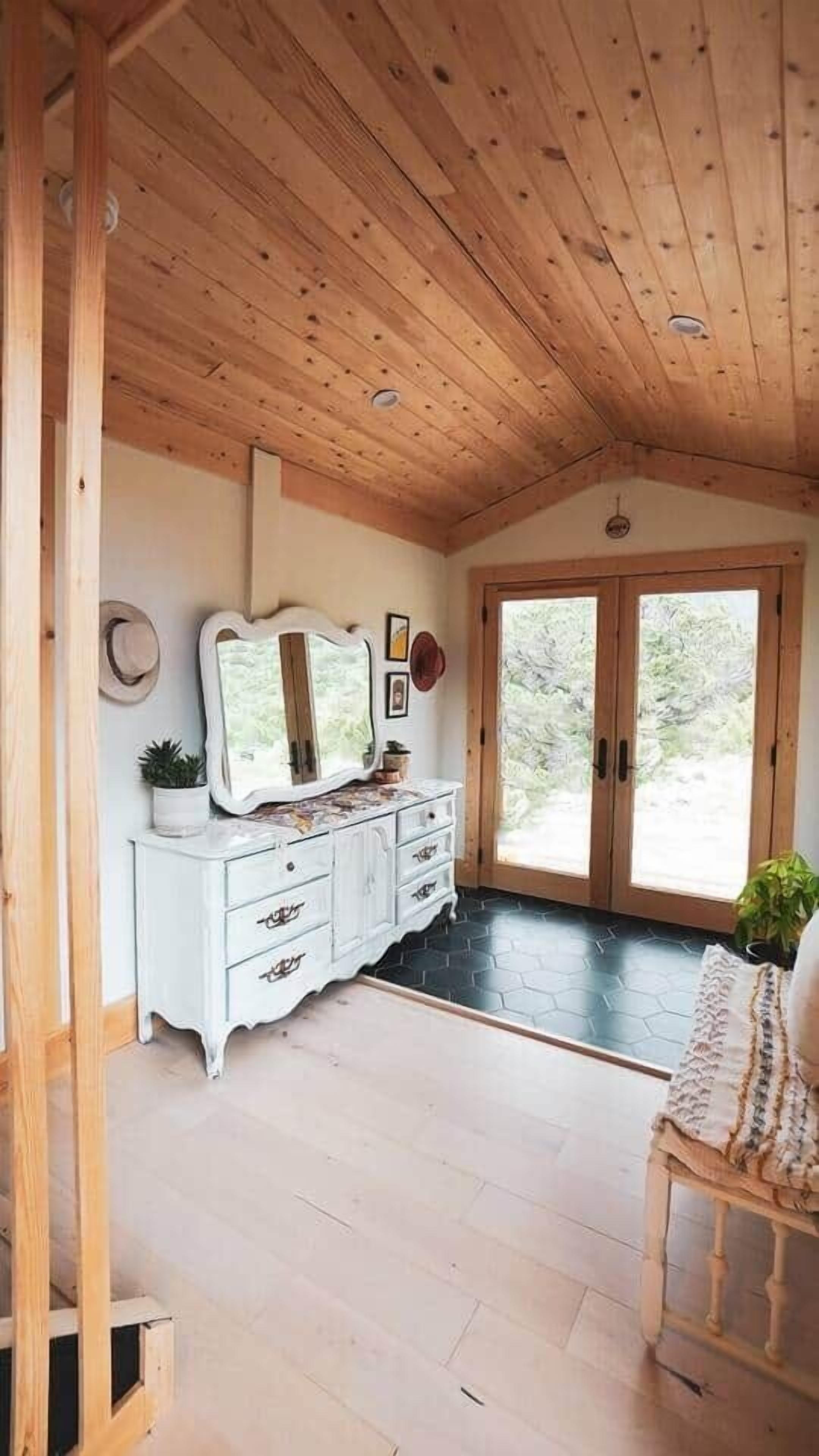 Habitación con techo de madera, un aparador blanco con cajones, un espejo y una puerta que da al exterior.