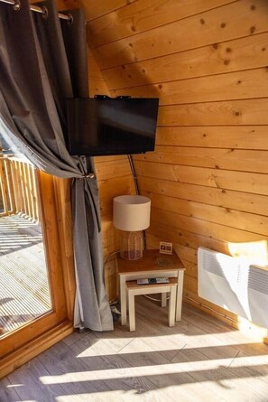 Ein gemütliches Zimmer mit Holzboden, einem an der Wand befestigten Fernseher, einem kleinen Tisch mit Lampe und einem Fenster mit Vorhängen.