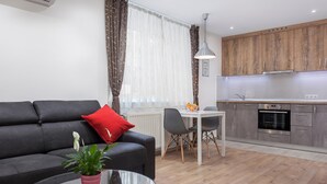 Ein modernes Wohnzimmer mit einer schwarzen Couch, einem kleinen Tisch mit einer Pflanze und einem Essbereich mit weißem Tisch und Stühlen. Im Hintergrund ist eine Küche mit Holzausstattung und Edelstahlgeräten zu sehen.