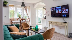 Una sala de estar con un sofá verde, sillas de madera, una mesa de comedor con mantel floral y un televisor que muestra varios canales.