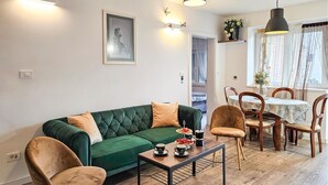 Una sala de estar con un sofá verde, dos sillones, una mesa de centro con tazas y un pastel, y un comedor con una mesa y sillas.