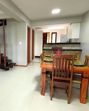 Un coin repas avec une table et des chaises en bois, une cuisine à l’arrière-plan et un escalier menant à un autre étage.
