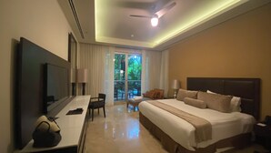 A hotel room with a large bed, a desk with a TV, a chair, and a balcony with a view of greenery. - 1BR suite, living room and kitchen in Vidanta Riviera Maya (Playa del Carmen)