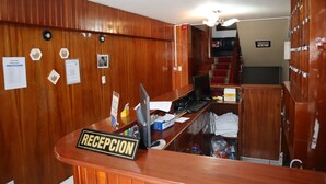 Et receptionsområde med en træreception, skiltet "RECEPCION", en computer samt diverse papirer og opslag på væggen.