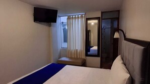 Et hotelværelse med en seng, et fjernsyn ophængt på væggen, et vindue med gardiner og et spejl.