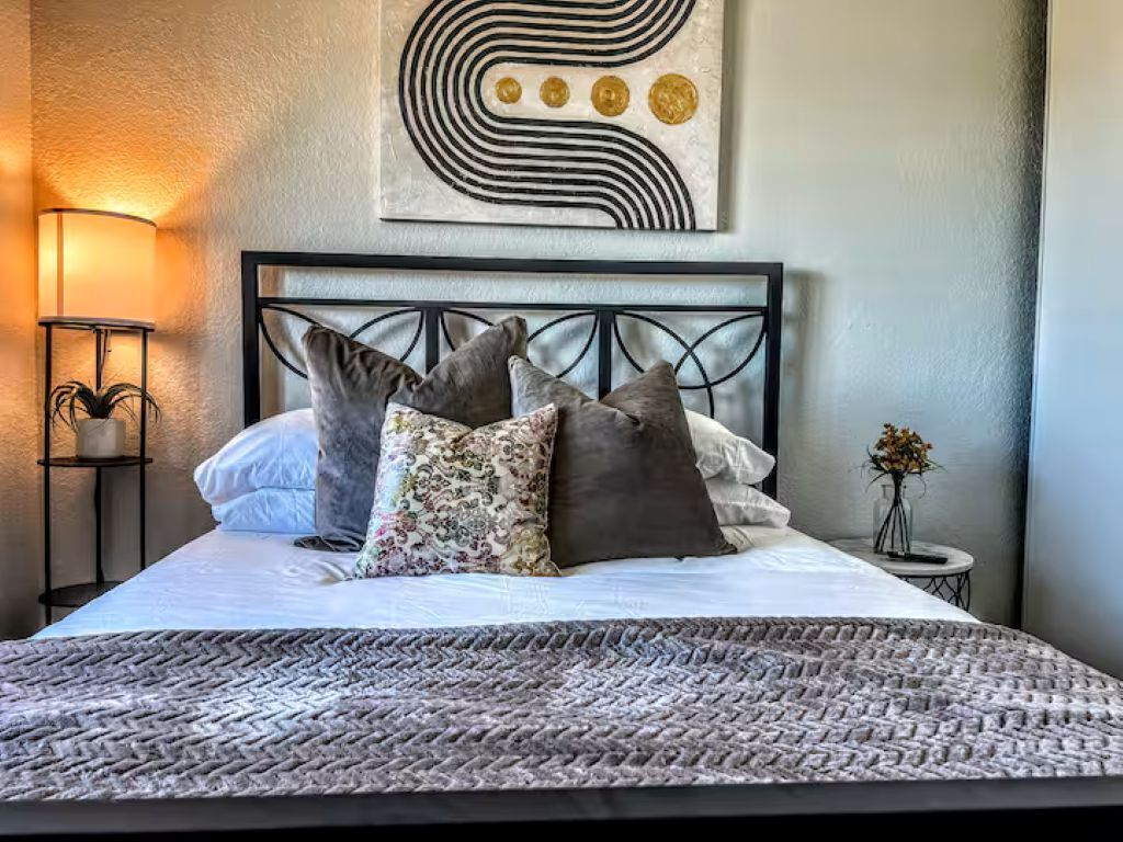 Un lit bien fait, avec une couvre-lit à motifs et plusieurs oreillers, une lampe de chevet et une œuvre d’art abstraite accrochée au mur.