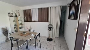 Una zona de menjador amb taula i cadires, un ventilador d'estàtua i una finestra amb cortines.