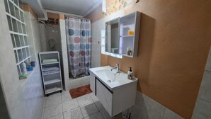 Un bany amb un lavabo blanc, un mirall, una petita estant amb articles de tocador i una zona de dutxa amb cortina estampada.