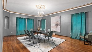 Uma sala de jantar com uma mesa posta para seis pessoas, rodeada por cadeiras, um lustre, cortinas cor de azul-turquesa e um grande quadro abstrato na parede.