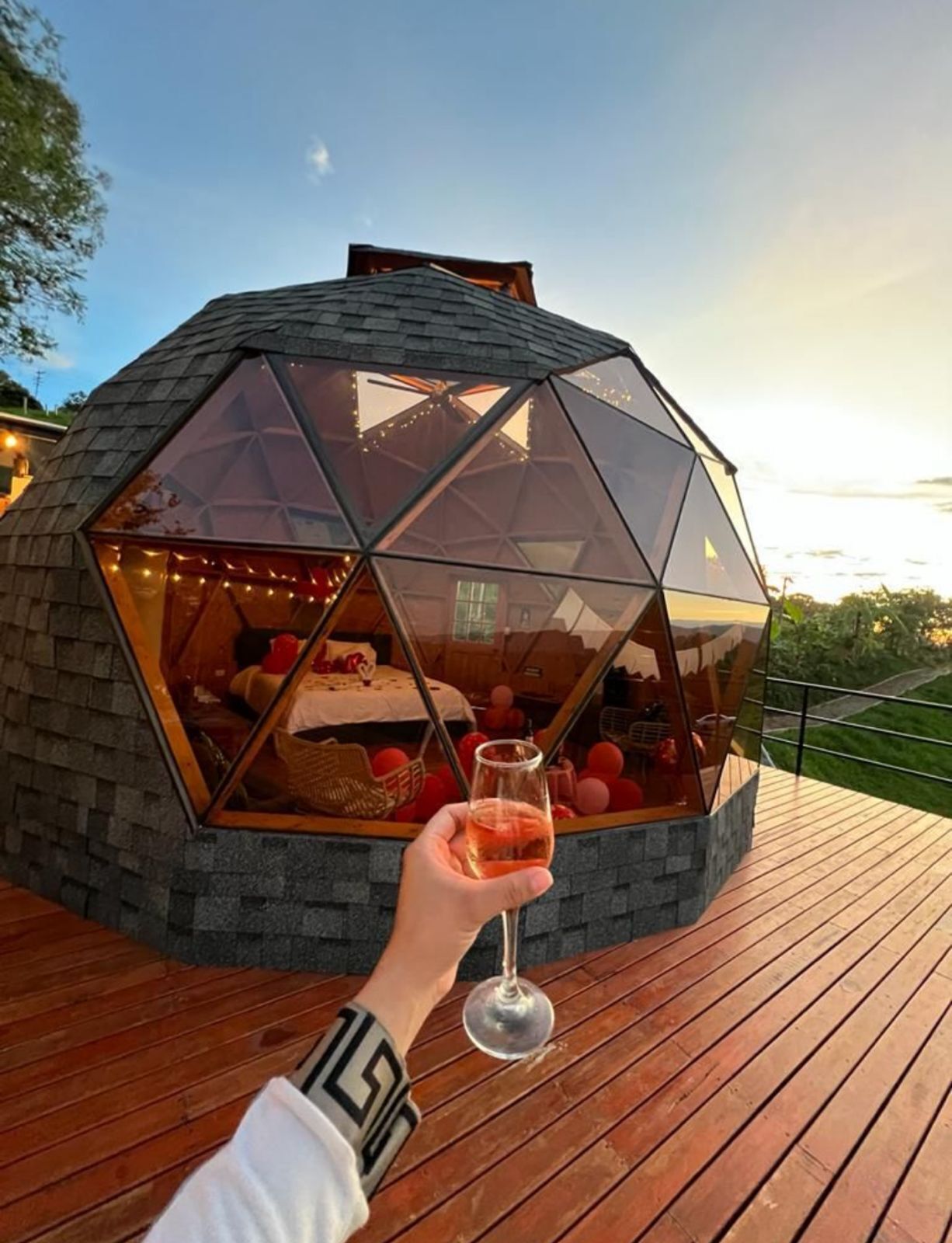 A geodesic dome structure with a transparent window, a wooden deck, and a person holding a glass.