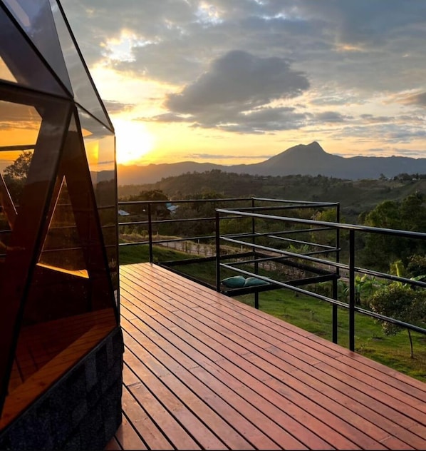 Une terrasse en bois avec une rambarde métallique, d’où l’on peut admirer un paysage montagneux au coucher du soleil.