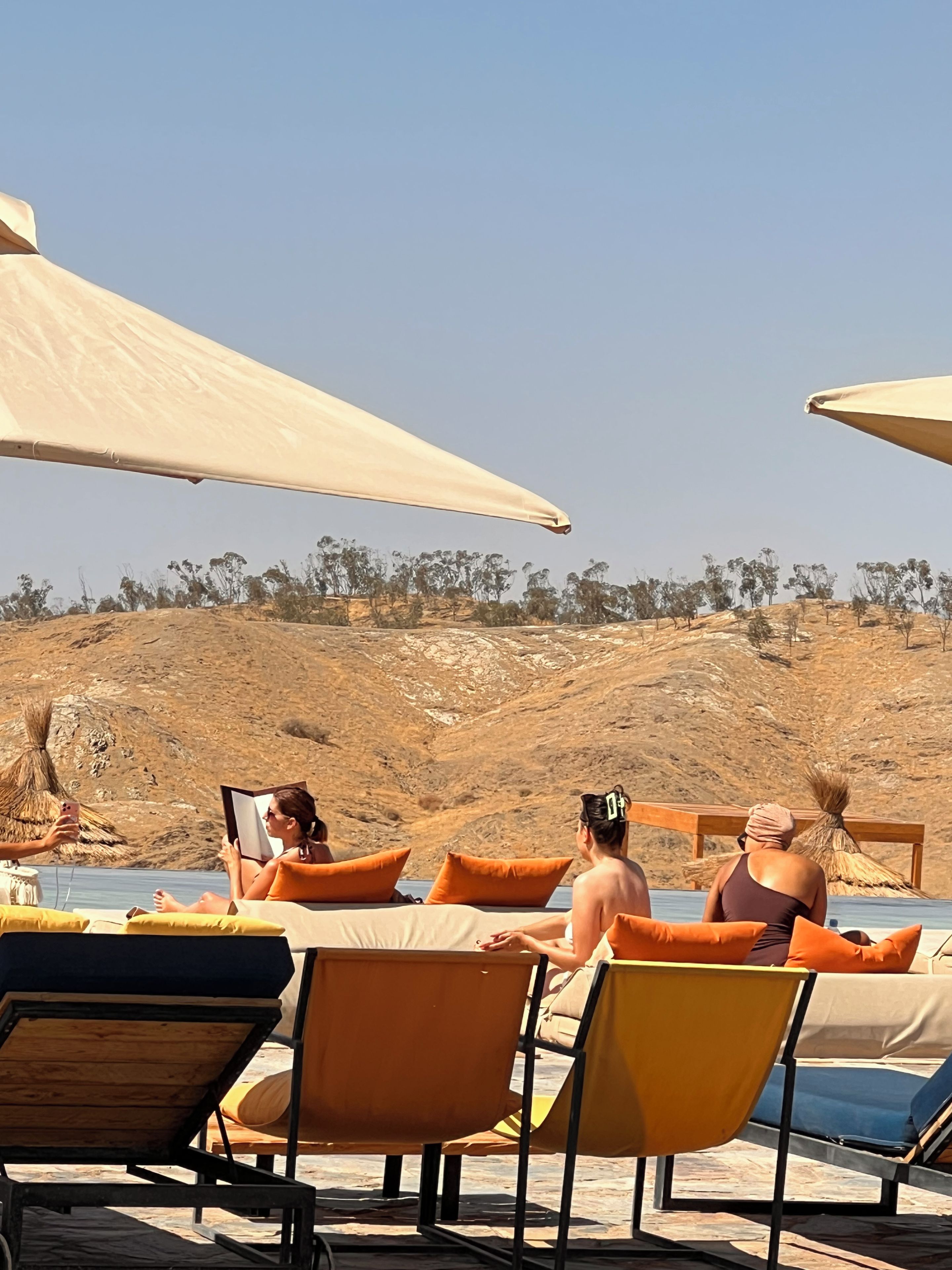 Zona junto a la piscina con sillones de descanso y sombrillas, cielo despejado y un paisaje montañoso al fondo.