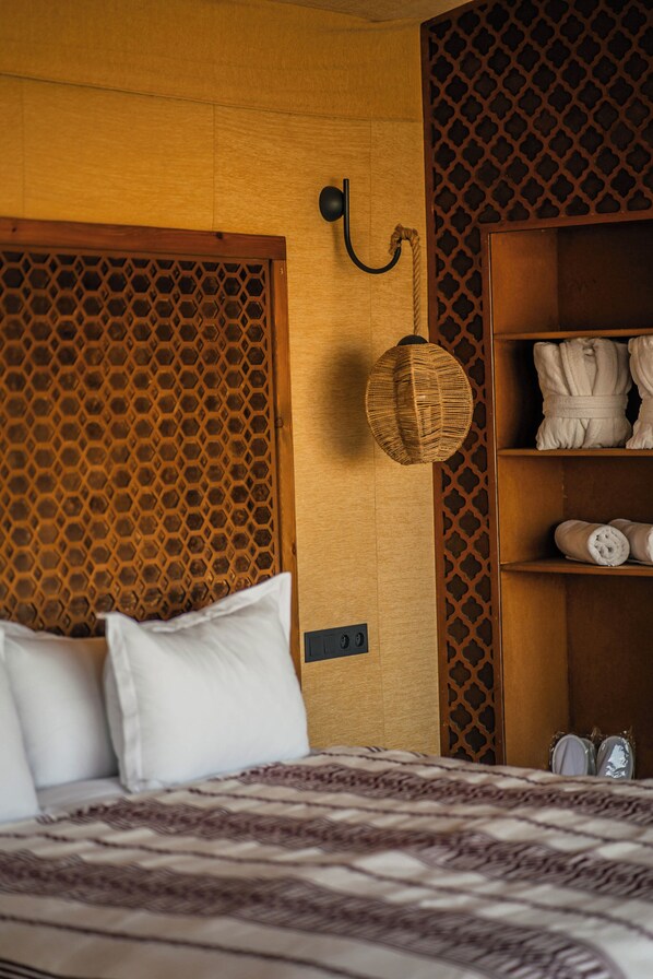 Une chambre à coucher avec un lit, des oreillers, un luminaire fixé au mur et une étagère en bois où sont posées des serviettes.