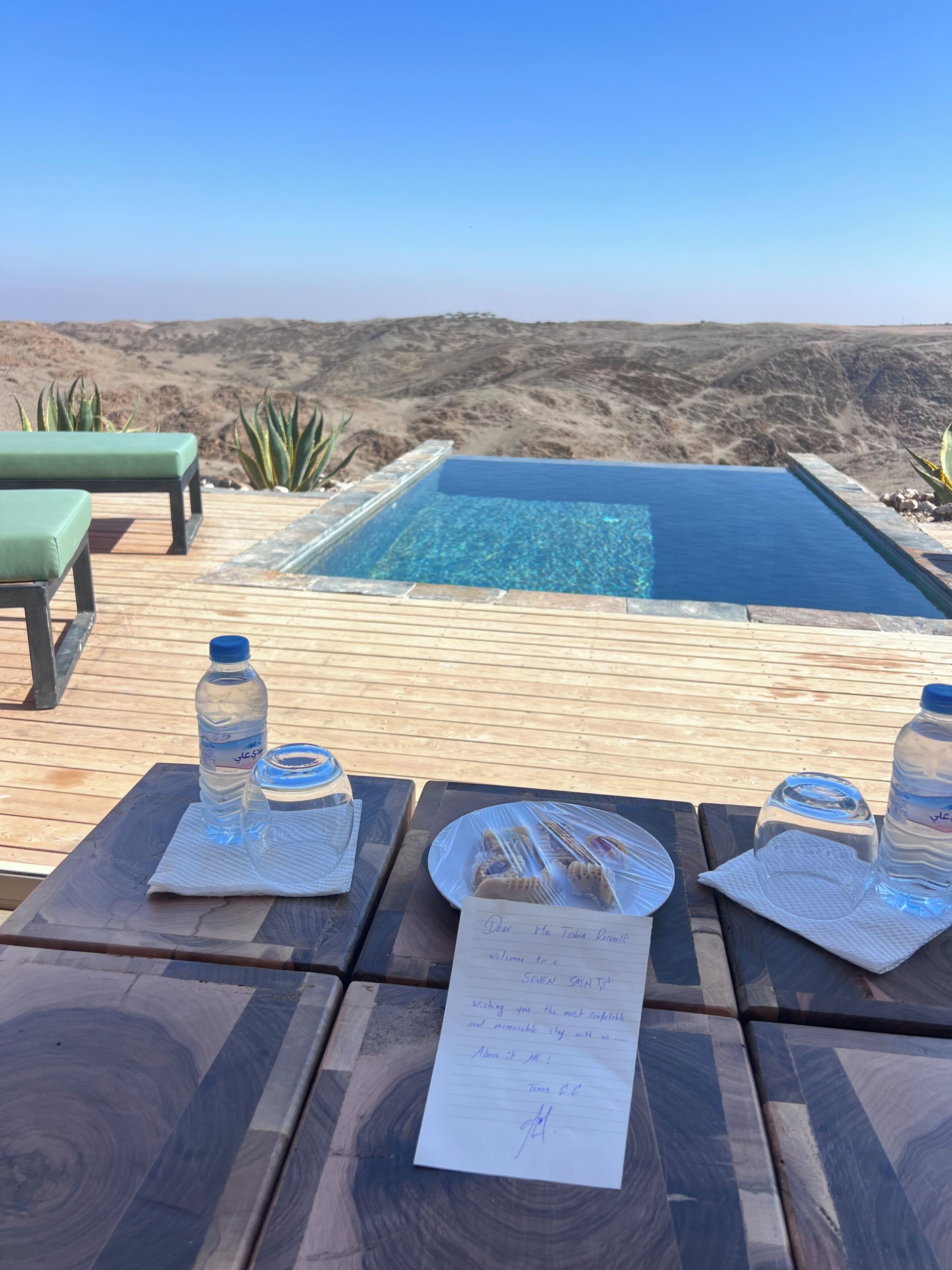 Ein Außenpoolbereich mit klarem, blauem Wasser, umgeben von einer Holzterrasse. Liegestühle und Tische sind mit Wasserflaschen und einem handschriftlichen Zettel bestückt.