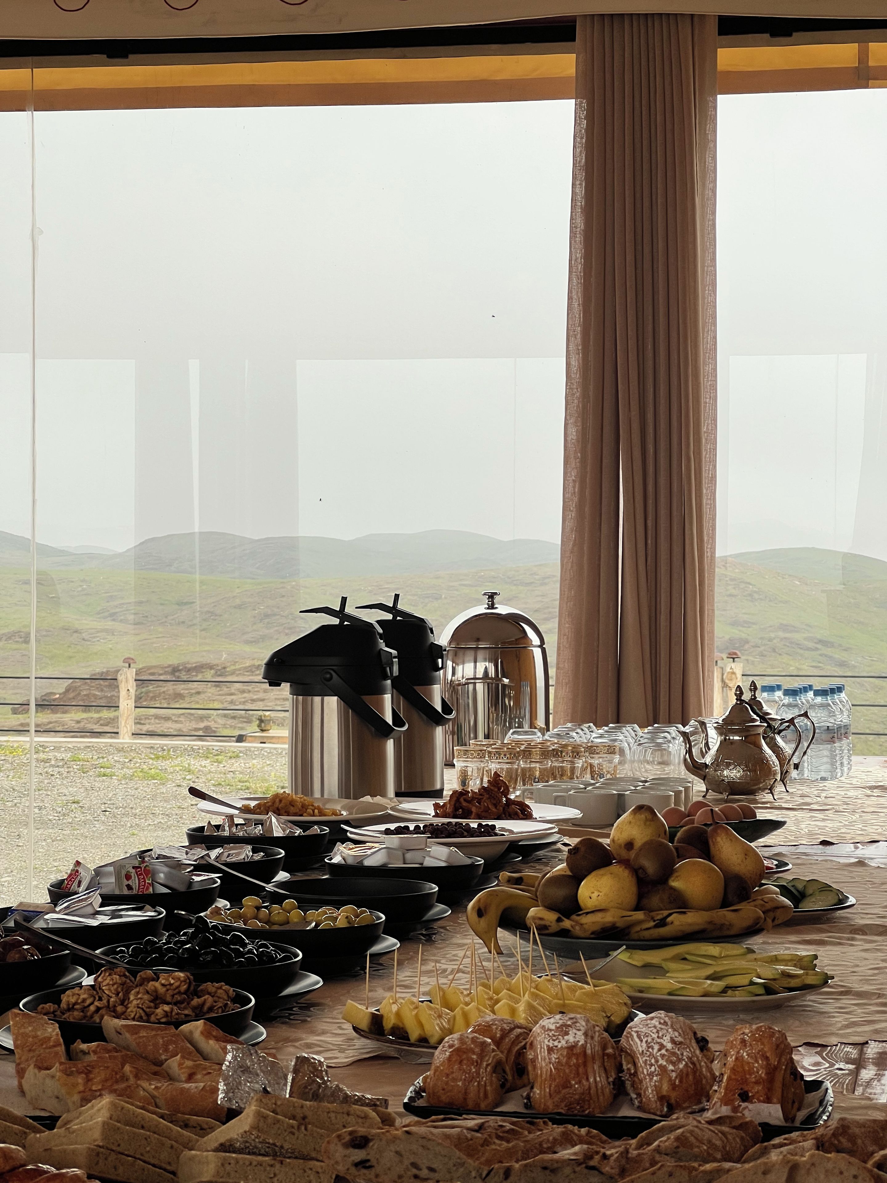 A buffet table with various food items and a view of a mountainous landscape through a window.