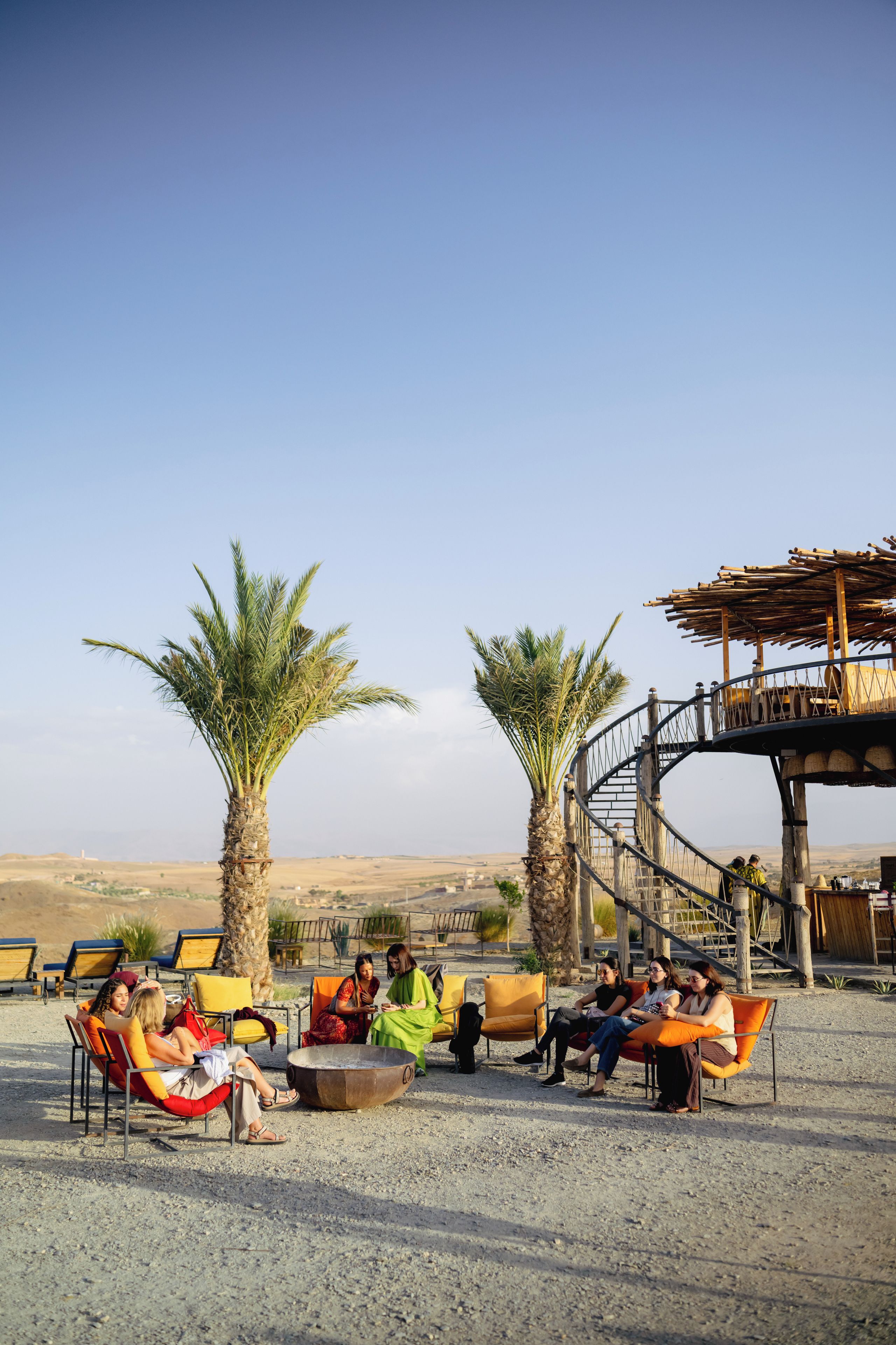 Un grupo de personas descansando en muebles de exterior, con palmeras y una estructura de madera al fondo.