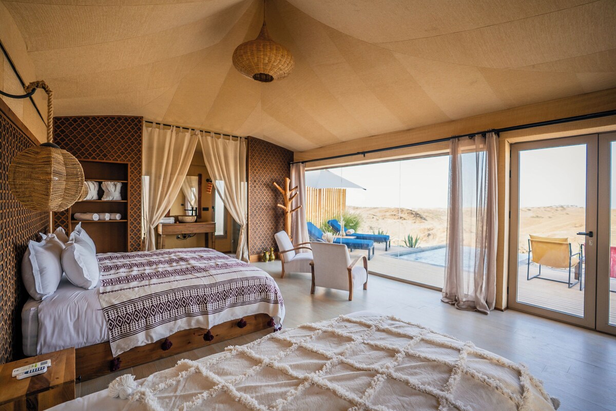 Een slaapkamer met een groot bed, een houten hoofdbord, een nachtkastje, een stoel en uitzicht op de woestijn door grote ramen.