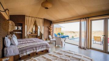 Ein Schlafzimmer mit einem großen Bett, einem Holzkopfende, einem Nachttisch, einem Sessel und Blick auf die Wüste durch große Fenster.