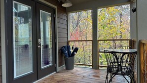 A covered porch with a wooden floor, a metal table and chair, and a view of autumn trees through large windows. - 2 Homes Mentone Getaway | Riverfront & Near Parks (Mentone)