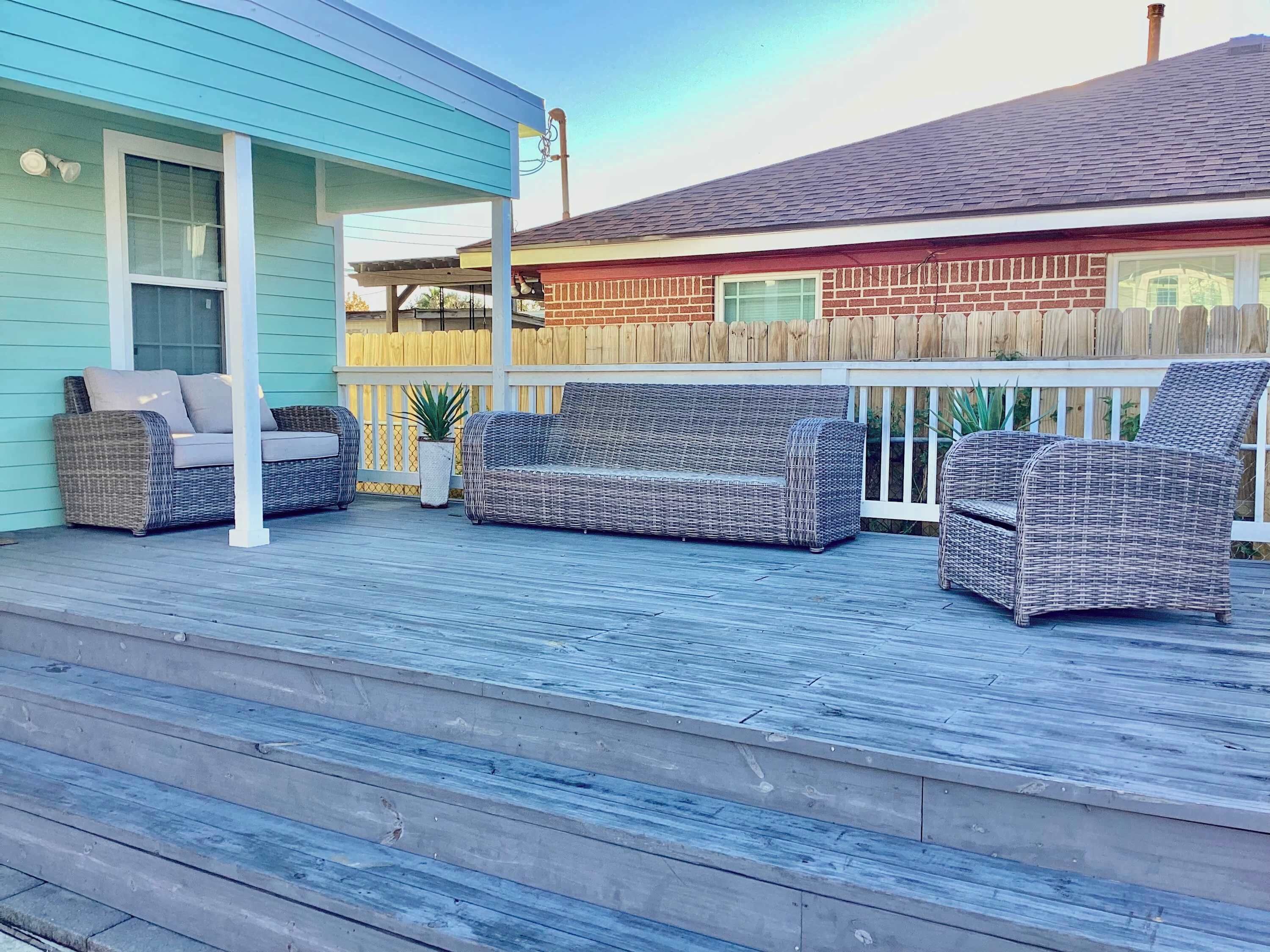 Una terraza techada al aire libre con mobiliario de mimbre, un edificio azul y una valla de madera.