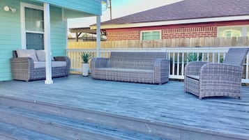 Una terraza techada al aire libre con mobiliario de mimbre, un edificio azul y una valla de madera.