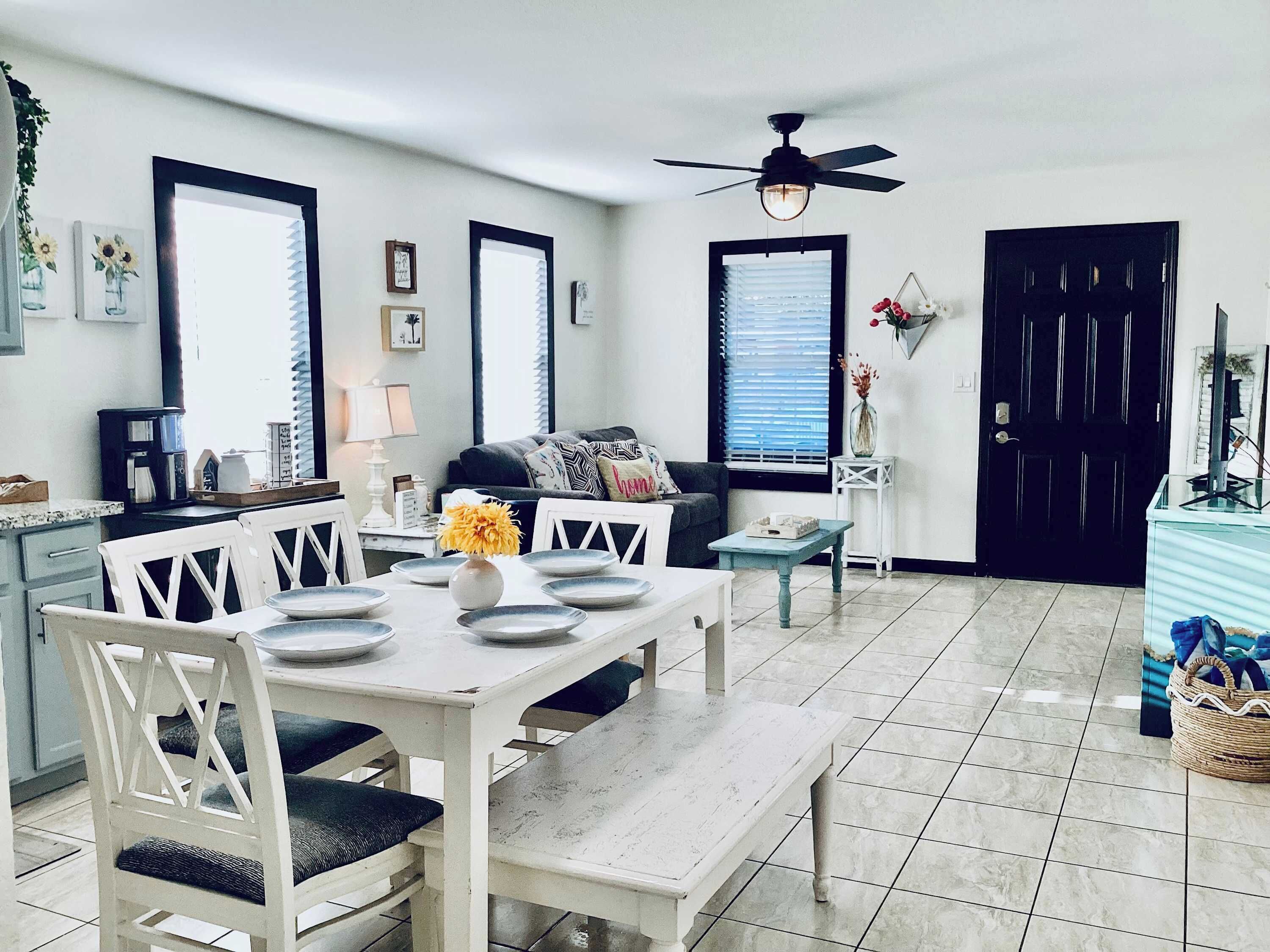 Comedor con una mesa blanca dispuesta para cuatro, un ventilador de techo y una puerta negra al fondo.