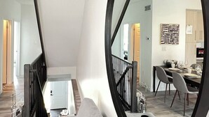 A modern living room with a large round mirror, a staircase, and a dining area with a table and chairs. - Bentley Commons Luxury 3 Bedroom Townhome in the Fallsview Tourist District (Niagara Falls)