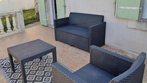 Un espace extérieur aménagé avec un canapé en osier foncé et deux tables assorties, un tapis à motifs et une porte verte en arrière-plan.