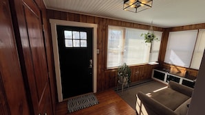 Una habitación con puerta de madera, ventana de marco negro, planta en maceta, sofá y lámpara en el techo.