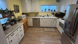 Una cocina con armarios blancos, un refrigerador de acero inoxidable, un microondas y un lavavajillas. Hay plantas en macetas en el alféizar de la ventana y un suelo de madera.