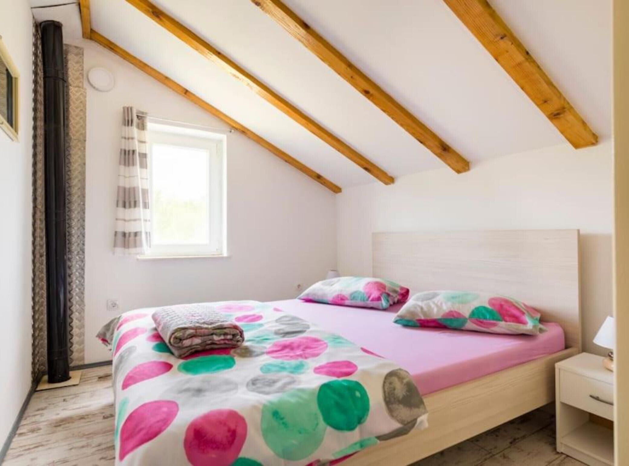 A bedroom with a wooden ceiling, a bed with colorful polka dot bedding, a bedside table, and a window with curtains.