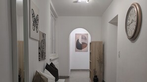 En gang med en træbænk, et lille bord og et tæppe. Malerier hænger på væggene, og der er et ur til højre.