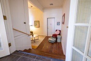 Um corredor com piso de madeira, uma cadeira vermelha, uma pequena mesa com abajur e uma porta com moldura.